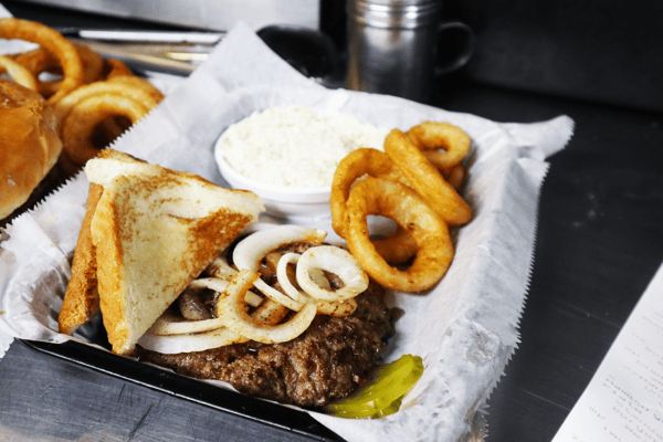 Hangry's Grill's juicy burger topped with grilled onions and toast, served with onion rings and a side of creamy sauce.
