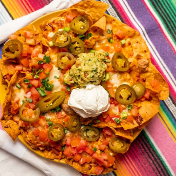 Nachos with melted cheese, diced tomatoes, sliced jalapeños, guacamole, and sour cream on a yellow plate.