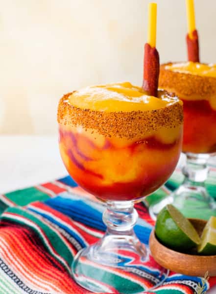 A mango chamoyada in a margarita glass with a chili rim. A tamarind straw and lime wedges are visible.