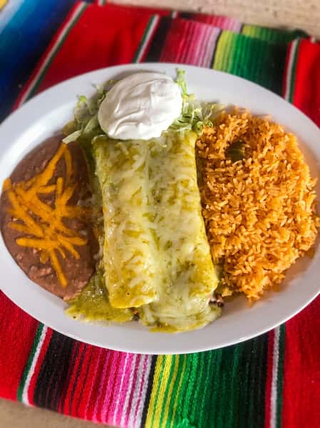 A plate of two green enchiladas with melted cheese, refried beans with shredded cheese, and Spanish rice.