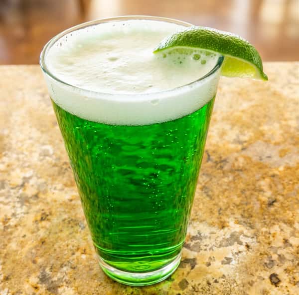Green beverage with white foam and a lime wedge in a tall glass, on a speckled countertop.