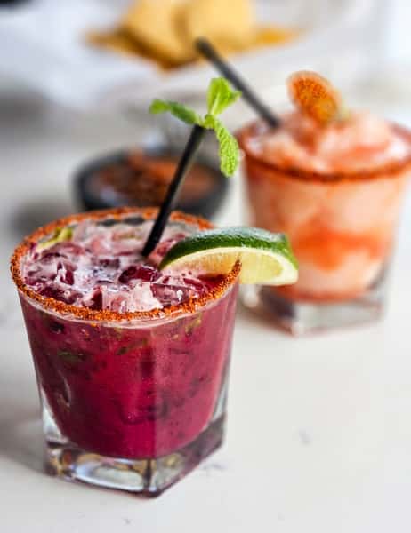 Close-up of a dark pink cocktail with a chili-lime rim, lime wedge, and mint, with a blurred lighter pink drink behind it.