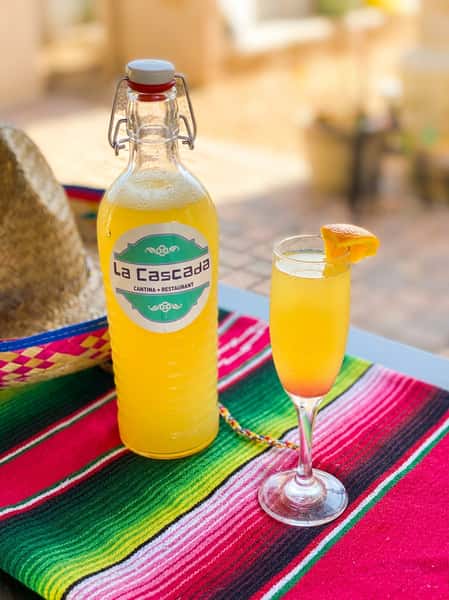 A glass bottle labeled "La Cascada" filled with yellow liquid, next to a mimosa with an orange slice, on a striped Mexican blanket.