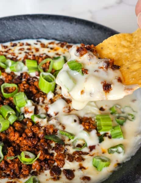 Queso dip with ground meat, minced green onions, and a tortilla chip scooping cheese.
