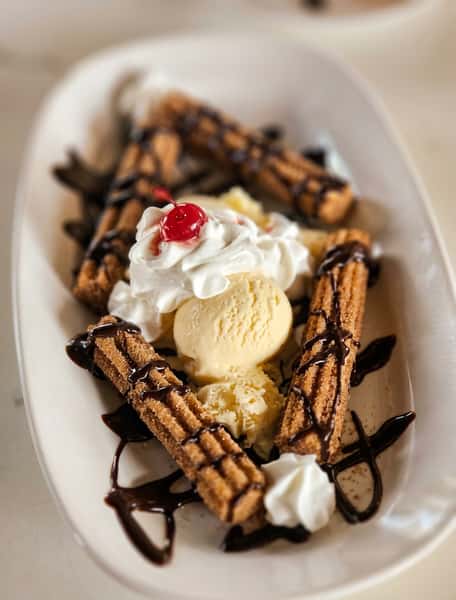 Churros topped with vanilla ice cream, whipped cream, a cherry, and chocolate drizzle on a white dish.