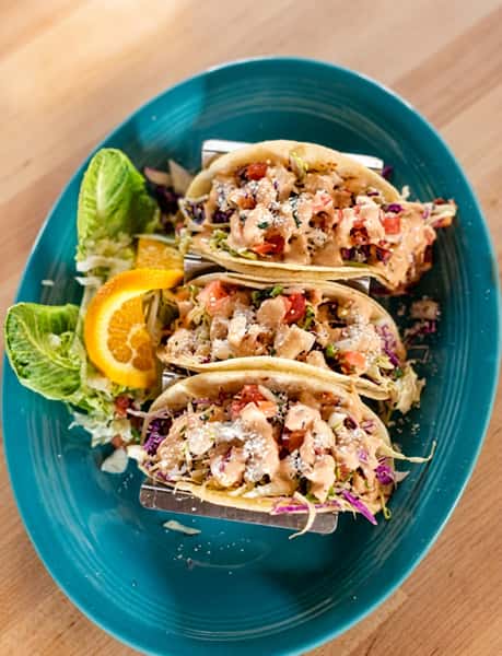 Three chicken street tacos in soft tortillas topped with sauce, micro-greens, and diced tomatoes, served with lettuce and an orange slice on a blue oval plate.