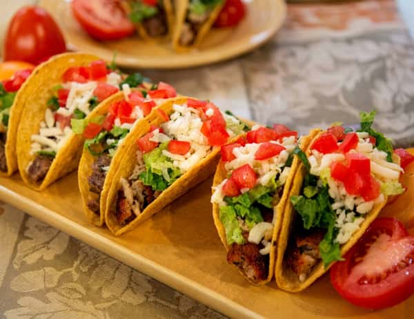 Five hard-shell tacos filled with meat, lettuce, shredded cheese, and diced tomatoes on a wooden platter.