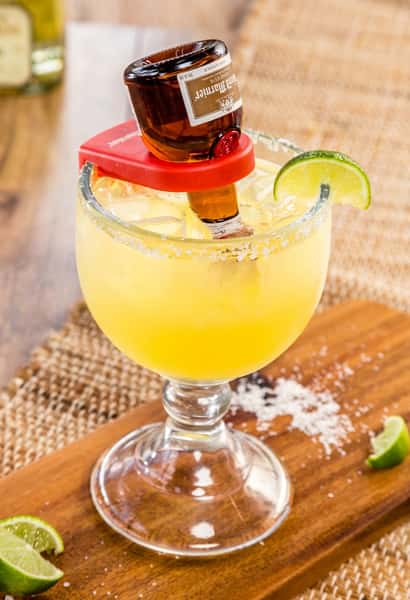 Margarita cocktail with a Grand Marnier bottle inverted into it, garnished with a lime slice, on a wooden board.