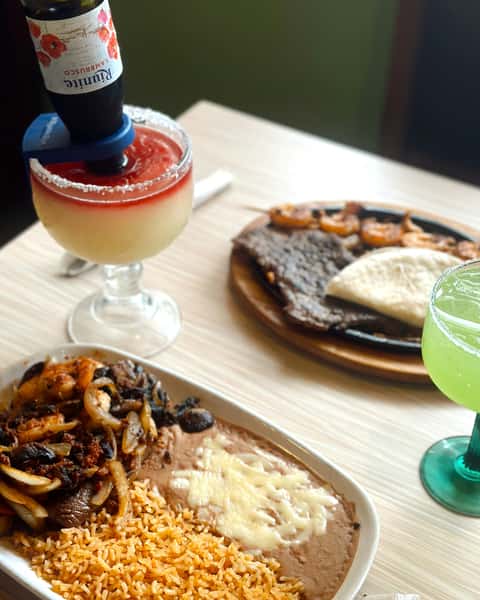 Mexican combo plate with grilled steak, sautéed shrimp, onions, mushrooms, refried beans topped with melted cheese, and seasoned rice, served alongside a sizzling fajita platter and warm flour tortillas. The table features a vibrant green margarita and a layered frozen margarita topped with an upside-down Tamarindo-flavored beer bottle for a bold twist.