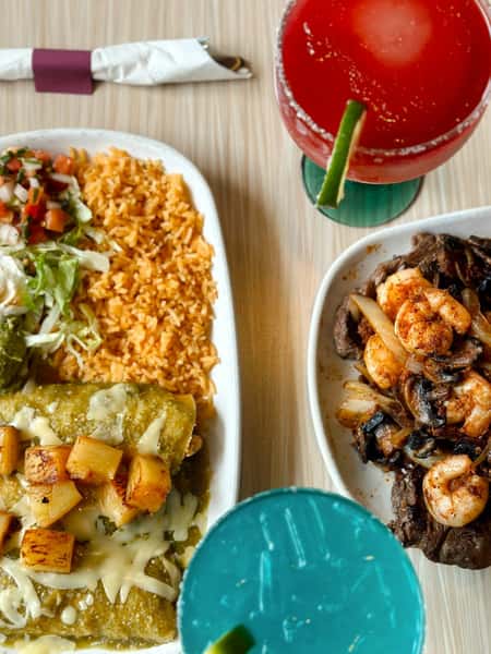 Colorful Mexican entrée featuring cheesy green enchiladas topped with grilled pineapple, served with Mexican rice, lettuce, sour cream, and pico de gallo, alongside a sizzling shrimp and steak fajita plate. Two vibrant margaritas—one red and one electric blue—add a festive touch to the table.