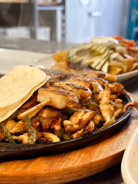 Sizzling chicken and shrimp fajitas served on a hot cast iron skillet with grilled onions and bell peppers, accompanied by warm flour tortillas. A loaded plate of crispy flautas topped with lettuce, sour cream, and avocado is visible in the background, capturing the vibrant flavors of authentic Mexican cuisine.