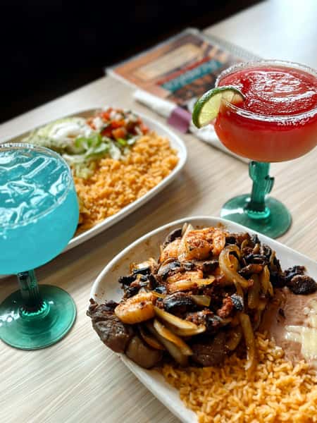 Colorful Mexican feast featuring a sizzling steak and shrimp fajita platter with grilled onions, mushrooms, and Mexican rice, paired with two vibrant margaritas—one blue and one red with a lime garnish. A second plate of seasoned rice with lettuce, sour cream, and pico de gallo is visible in the background, set on a casual restaurant table.