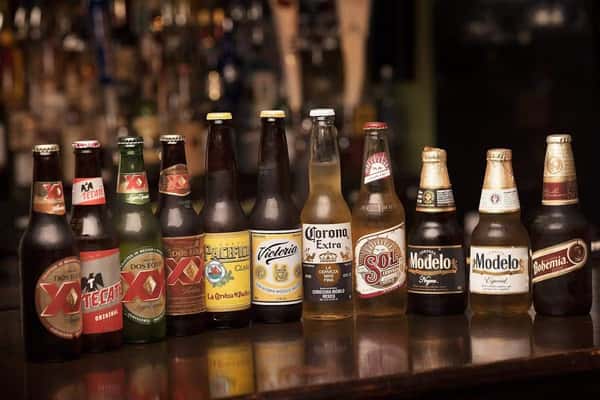 Assorted lineup of popular Mexican beers including Dos Equis, Tecate, Pacifico, Victoria, Corona Extra, Sol, Modelo, and Bohemia, displayed on a bar counter with a blurred background of taps and spirits, showcasing authentic Mexican beverage options.