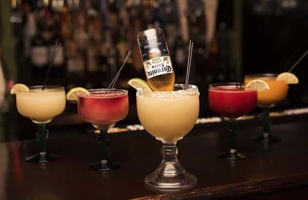 Assortment of colorful frozen margaritas garnished with lime wedges on a bar counter, featuring a standout Corona-Rita with an upside-down Corona beer bottle. The drinks come in vibrant hues of yellow, red, and orange, perfect for showcasing a lively Mexican bar atmosphere.