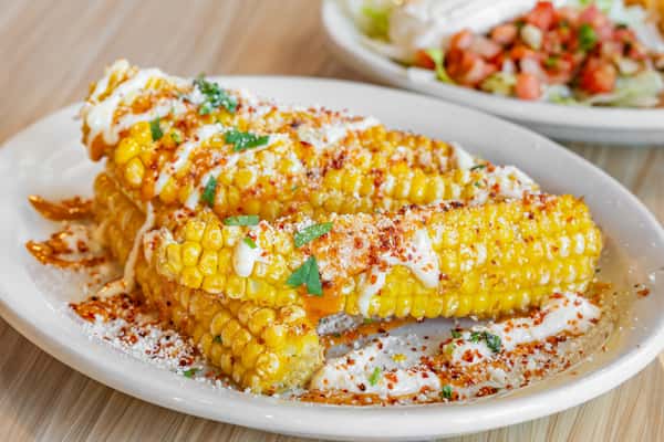 Grilled Mexican street corn topped with creamy mayo, crumbled cotija cheese, chili powder, and chopped cilantro, served on a white plate.