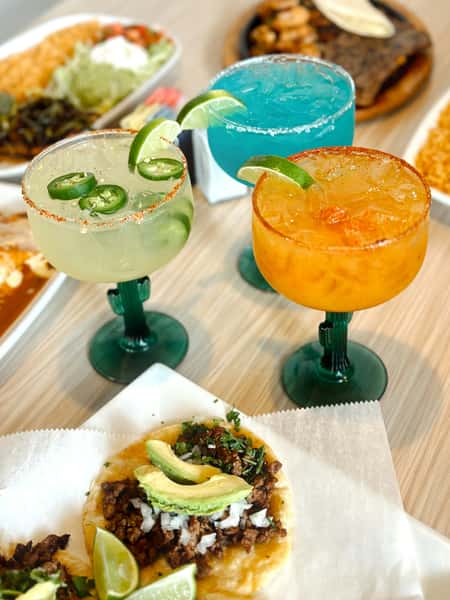 Vibrant Mexican meal with street-style tacos topped with avocado, onions, cilantro, and lime wedges, paired with colorful margaritas in lime, blue, and mango flavors. Plates of Mexican rice and sizzling fajitas are visible in the background, creating a festive dining scene.
