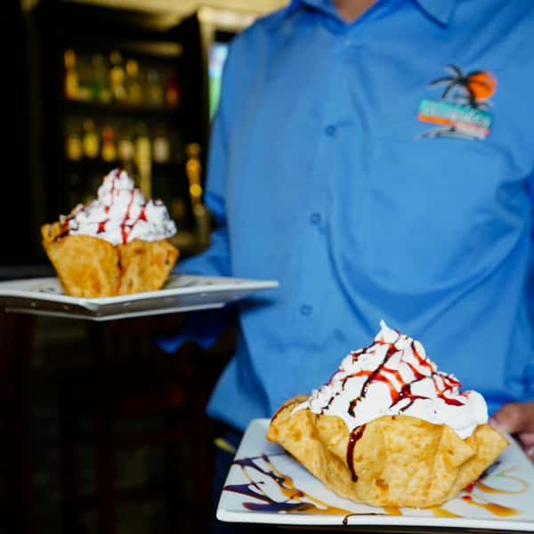 Server presenting two crispy fried tortilla dessert bowls filled with vanilla ice cream, topped with whipped cream and drizzled with chocolate and strawberry syrup, served on square plates in a festive Mexican restaurant setting.