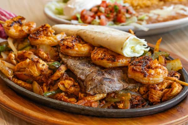 Sizzling surf and turf fajitas with grilled shrimp, steak, and chicken, served over sautéed peppers and onions on a cast iron skillet, with a warm flour tortilla on the side.