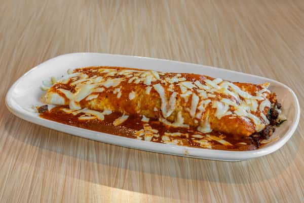 Large beef burrito topped with melted cheese and rich red enchilada sauce, served on a white plate on a wooden table.