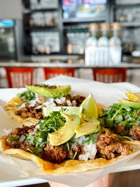 Freshly made street-style carne asada tacos topped with diced onions, cilantro, avocado slices, and lime wedges, served on soft corn tortillas in a lively Mexican restaurant bar setting.