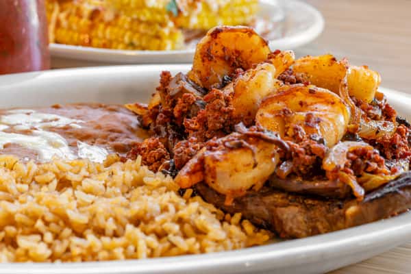 Grilled shrimp and crumbled chorizo sausage over a juicy steak, served with seasoned Mexican rice and refried beans topped with melted cheese on a white plate.