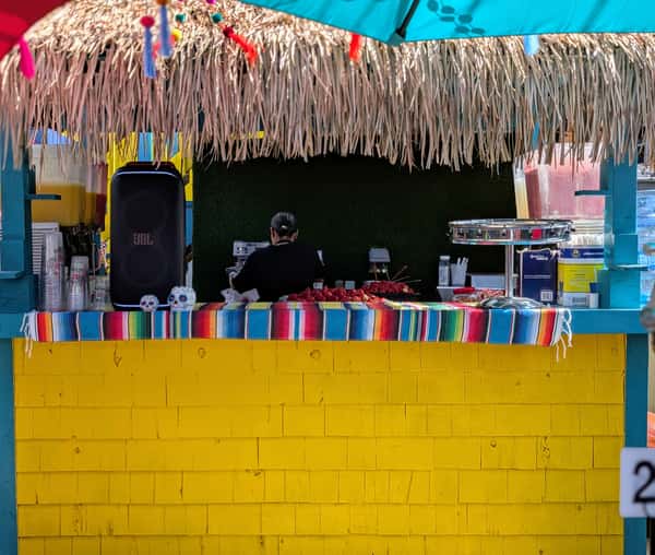 vendor prepares food at a brightly colored stand decorated with a thatched roof and colorful tablecloth