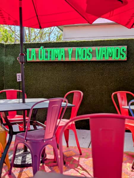 The words "La Última y Nos Vamos" are lit in neon, set against a green backdrop