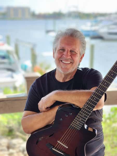 Grey hair, tan, musician holding electric guitar with Halifax river in the background