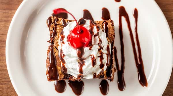 Chocolate brownie topped with whipped cream, a maraschino cherry, and chocolate sauce drizzle on a white plate.