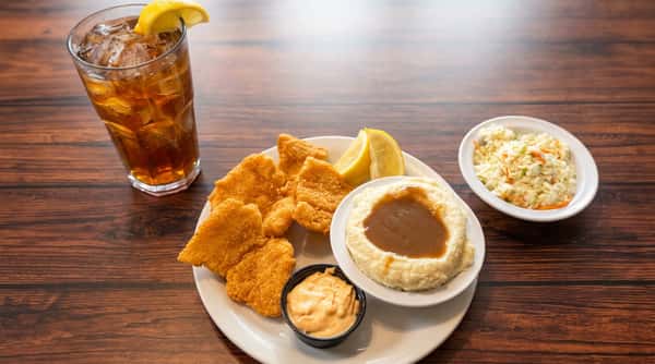 Fried fish platter with mashed potatoes and gravy, coleslaw, dipping sauce, and a Coca-Cola on a wooden table.