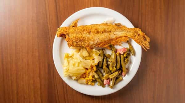 Fried fish fillet with sides of stewed cabbage, corn, and green beans with ham on a white plate.