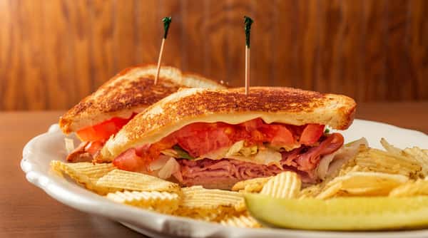Toasted sandwich with ham, lettuce, tomato, and cheese, served with ripple-cut potato chips and a pickle spear.