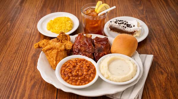 Platter with fried chicken, BBQ ribs, baked beans, mashed potatoes with gravy, roll, yellow corn, chocolate pie, and iced tea with lemon.