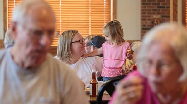 A woman with glasses looks at a young girl in a pink Paw Patrol bib with her hand on the woman's shoulder, indoors.