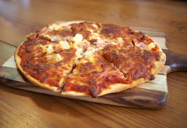 Golden-baked Hawaiian pizza with melted cheese, pineapple chunks, and ham on a thin crust, served on a rustic wooden paddle — great for showcasing tropical-inspired pizza menus.