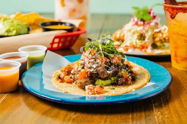 A vibrant Milagro Tacos Cantina spread featuring a loaded taco with meat, guacamole, and pico de gallo on a blue plate, alongside sauces and a drink.