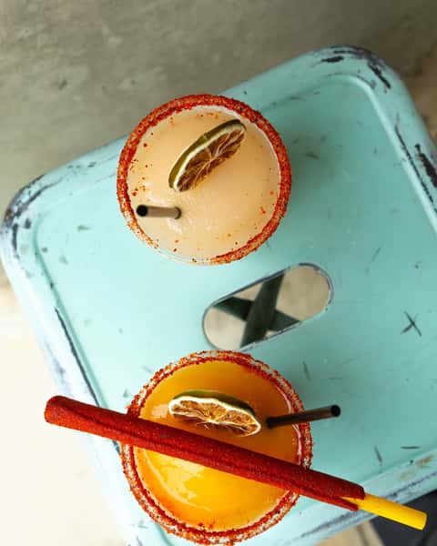 Two vibrant, chili-rimmed margaritas garnished with dried lime and a chili candy stick, served on a teal stool at Milagro Tacos Cantina.