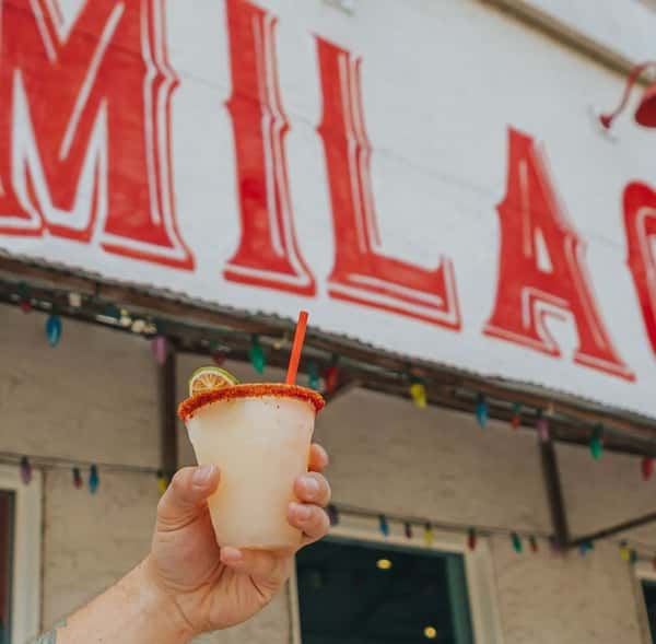 Hand holding a frosty Milagro Tacos Cantina margarita with a chili-lime rim, set against the vibrant red sign and colorful lights.