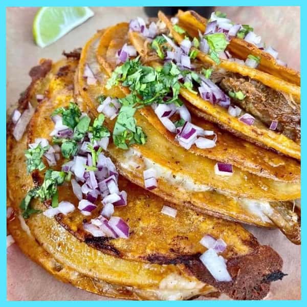 Close-up of golden-brown crispy tacos from Milagro Tacos Cantina, generously topped with red onions and cilantro, with a lime slice.