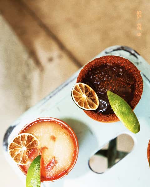 Two chili-rimmed margaritas with dried and fresh lime garnishes rest on a light blue surface at Milagro Tacos Cantina.