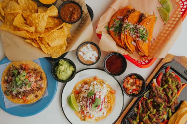 A colorful spread of Mexican food from Milagro Tacos Cantina, featuring birria tacos, chips, guacamole, fajitas, and a tostada.