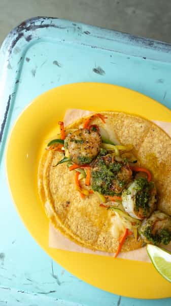Milagro Tacos Cantina's vibrant shrimp taco, topped with green sauce, fresh vegetables, and lime, served on a bright yellow plate against a rustic blue background.
