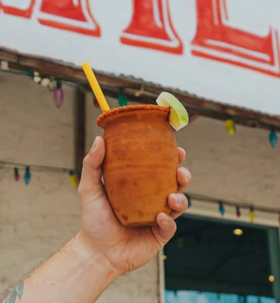 A hand holds a terracotta mug drink with a chili-lime rim, lime wedge, and yellow straw, against the Milagro Tacos Cantina background.