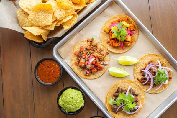 Milagro Tacos Cantina's vibrant spread of assorted tacos, tortilla chips, red salsa, and green guacamole on a wooden table.