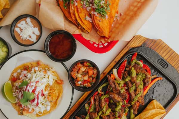 A colorful array of Milagro Tacos Cantina's Mexican dishes, featuring birria tacos, sizzling fajitas with peppers, and a loaded tostada, alongside various vibrant dips.