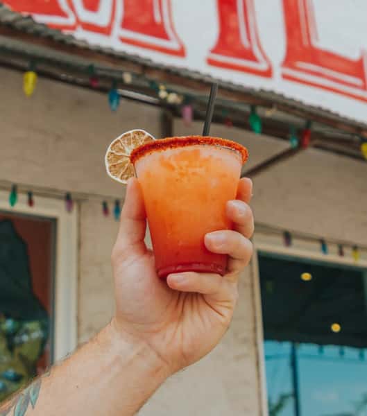 A hand holds a vibrant orange-red cocktail with a chili-lime rim and dried lime garnish, set against a background of festive lights at Milagro Tacos Cantina.