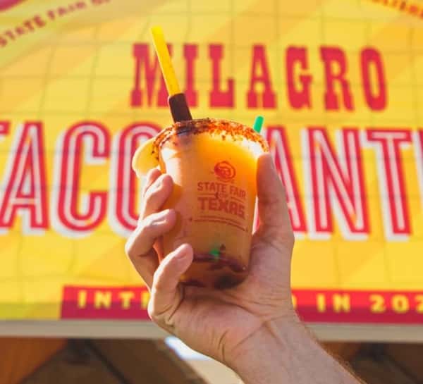 A hand holds a vibrant orange, chamoy-rimmed drink in a "State Fair of Texas" cup, against a bright yellow "Milagro Tacos Cantina" banner.