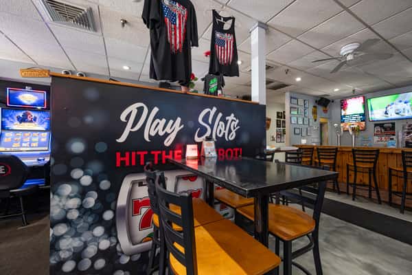 Casual gaming-themed dining area with high-top tables, chairs, and "Play Slots" signage, set against a backdrop of slot machines and a lively bar scen