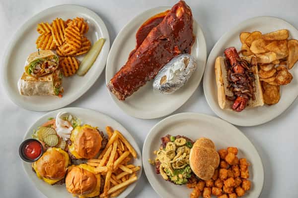 Assorted American comfort food platters featuring BBQ ribs with baked potato, a grilled sausage sandwich with chips, cheeseburger sliders with fries, a loaded burger with tater tots, a Southwest wrap with waffle fries, and more — a hearty spread perfect for casual dining or game day meals.