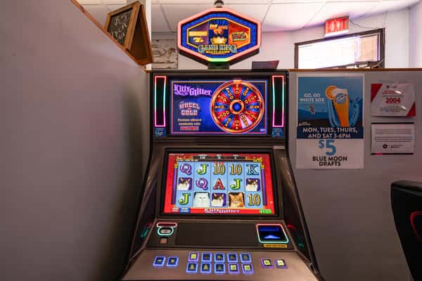Colorful slot machine featuring the "Kitty Glitter" game with a digital wheel and neon lights, set in a gaming corner with drink specials and promotional signage.